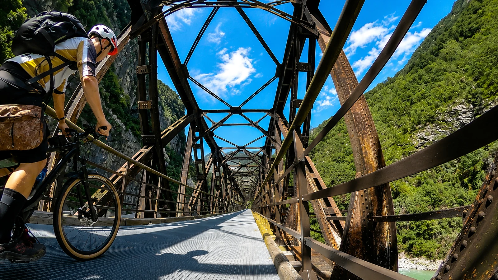 Ciclista su ponte metallico abbandonato in fondo valle, tra pareti rocciose e vegetazione rigogliosa.