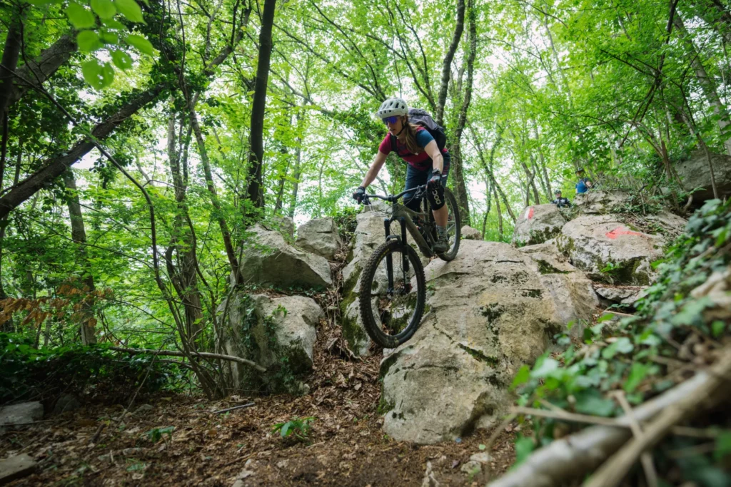 Ciclista in mountain bike percorre un sentiero tecnico tra grandi rocce e vegetazione fitta in una foresta verde e rigogliosa.