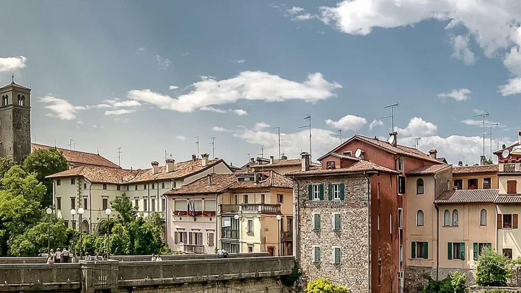 Centro storico medievale con edifici in pietra e mattoni, torre campanaria e ponte antico sotto cielo nuvolato.