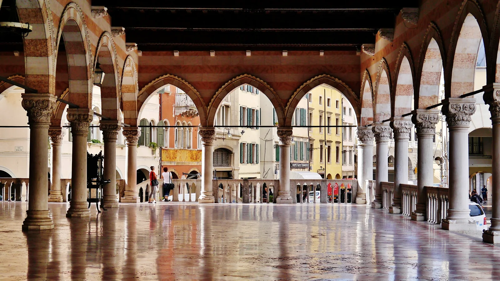 Loggia medievale con arcate a strisce rosse e bianche, pavimento bagnato riflettente, piazza storica sullo sfondo.