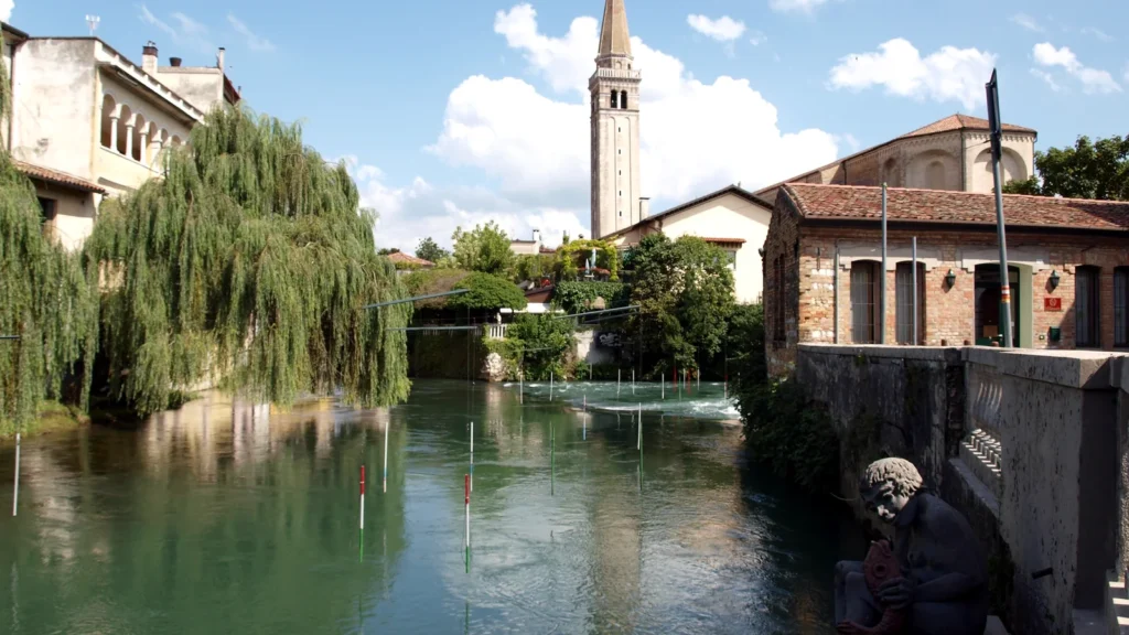 Canale con acqua verde circondato da edifici storici in mattoni e campanile sullo sfondo in una cittadina veneta.