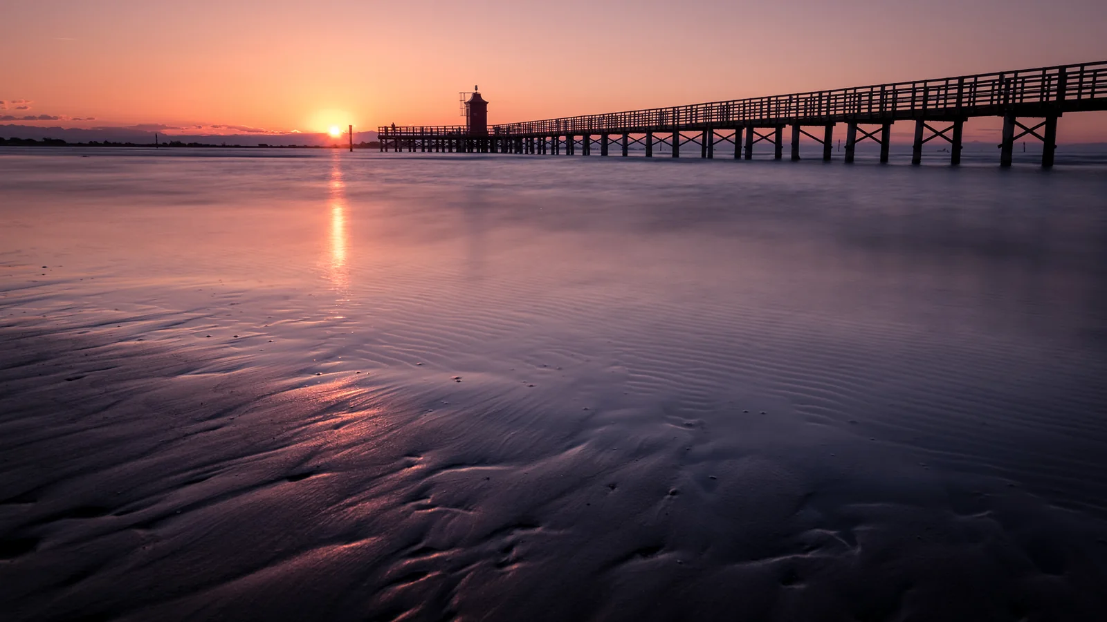 Ponte in legno al tramonto su spiaggia sabbiosa con faro sullo sfondo e cielo rosato.