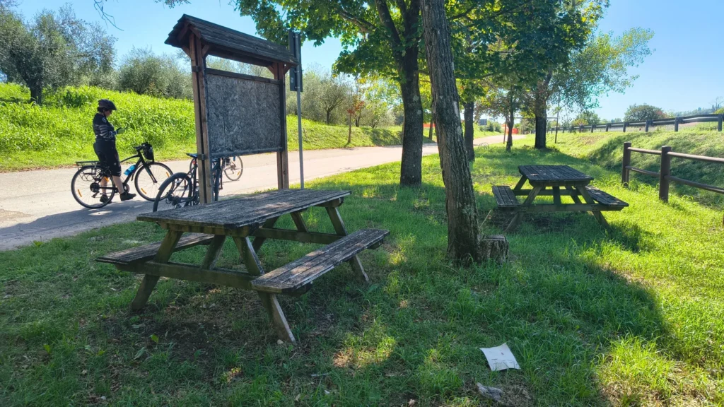 Ciclista con bici accanto a una pensilina in legno lungo una ciclovia, con tavoli da picnic sotto alberi frondosi.