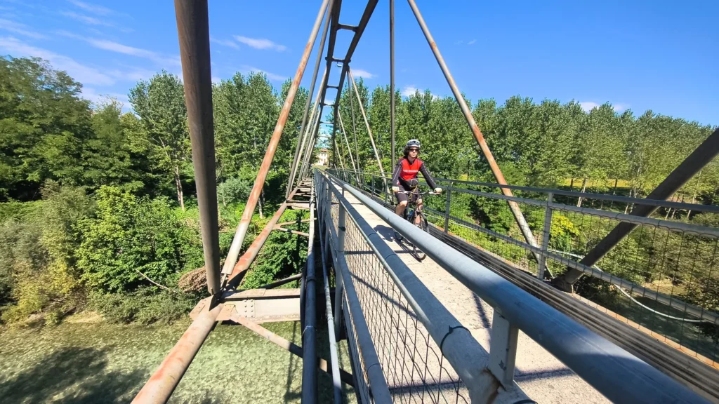 Ciclista in giubbotto rosso attraversa un ponte sospeso metallico sopra la vegetazione, bosco sullo sfondo.