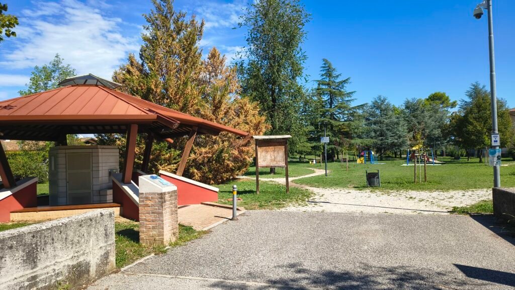 Area di sosta cicloturistica con pensilina rossa, parcheggio bici e verde circostante sotto cielo sereno.
