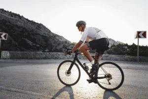 Ciclista in maglia bianca su strada in curva davanti a parete rocciosa grigia con cartelli di direzione.