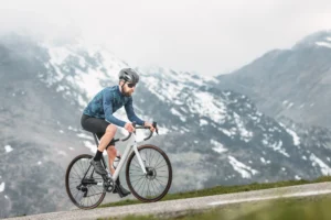 Ciclista su strada di montagna con bici da corsa bianca, sfondo di vette innevate.