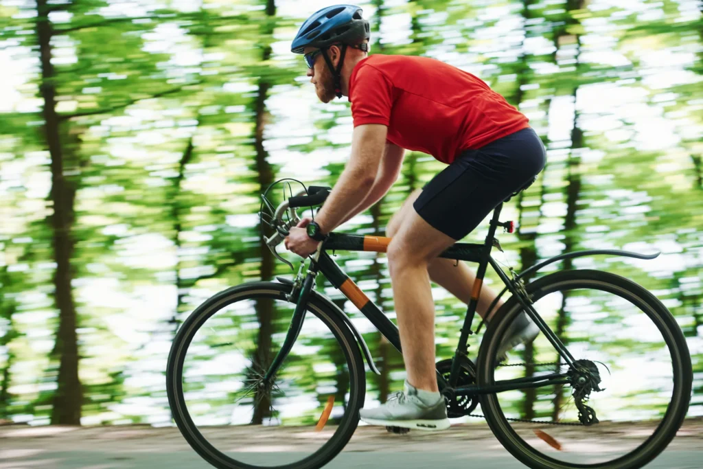 Ciclista in maglia rossa pedala veloce su strada tra boschi di conifere e faggi.