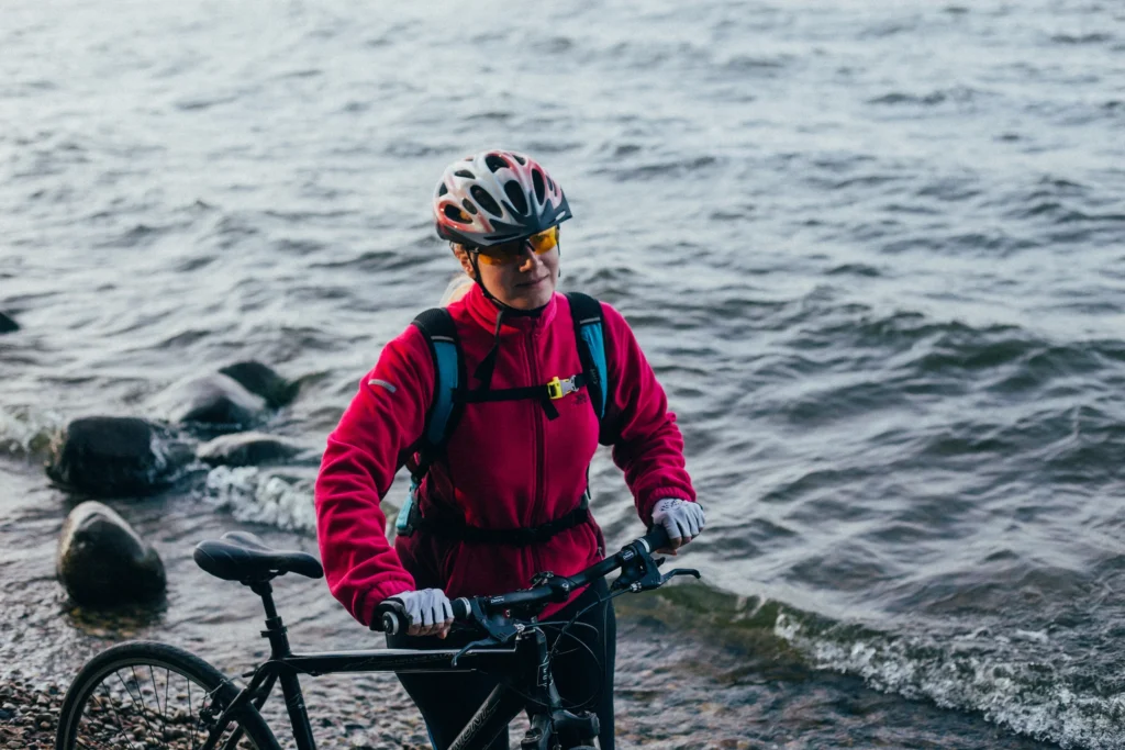 Ciclista in giacca rossa spinge la bici sulla spiaggia con il mare sullo sfondo.