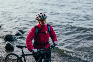 Ciclista in giacca rossa spinge la bici sulla spiaggia con il mare sullo sfondo.