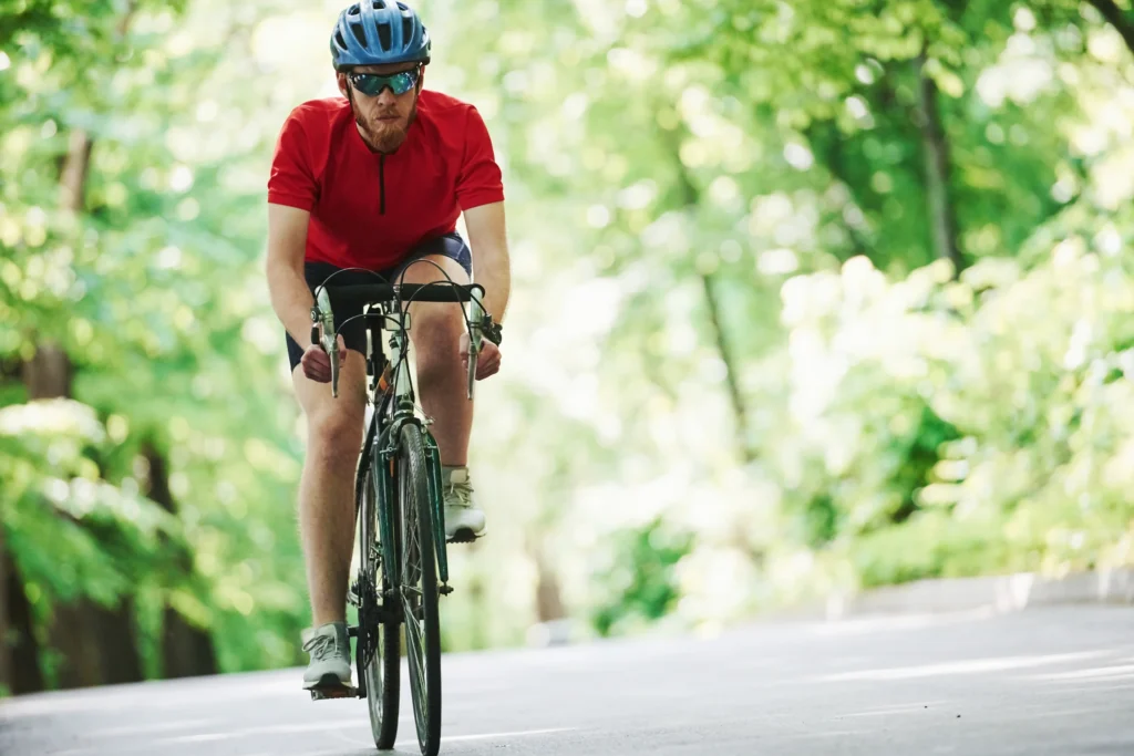 Ciclista in maglietta rossa pedala su strada asfaltata circondata da vegetazione verde lussureggiante.