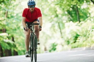 Ciclista in maglietta rossa pedala su strada asfaltata circondata da vegetazione verde lussureggiante.