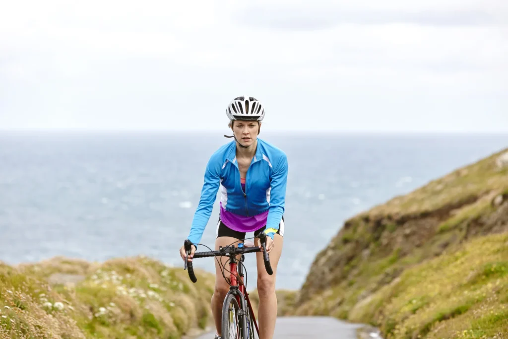 Ciclista in maglia blu percorre una strada costiera tra scogliere verdi e mare calmo sullo sfondo.