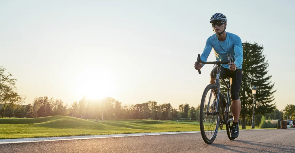 Ciclista in maglia azzurra pedala su una pista asfaltata al tramonto, con prati verdi e alberi sullo sfondo.