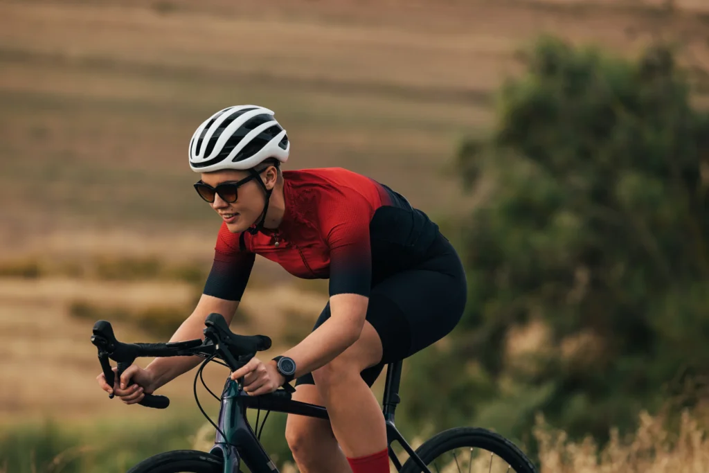 Ciclista in maglia rossa e nera pedala su strada collinare con paesaggio agricolo sfumato sullo sfondo.