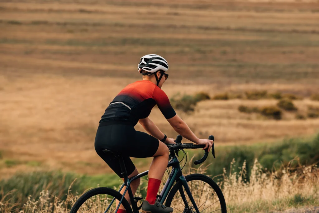Ciclista in maglia rossa e nera in sella su strada rurale con campi ondulati sullo sfondo.