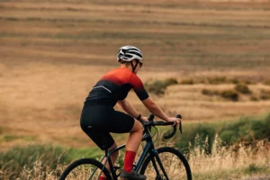 Ciclista in maglia rossa e nera in sella su strada rurale con campi ondulati sullo sfondo.