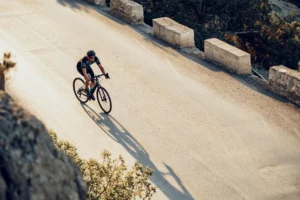 Ciclista in maglia nera su schotterweg bianco circondato da blocchi di cemento e vegetazione.