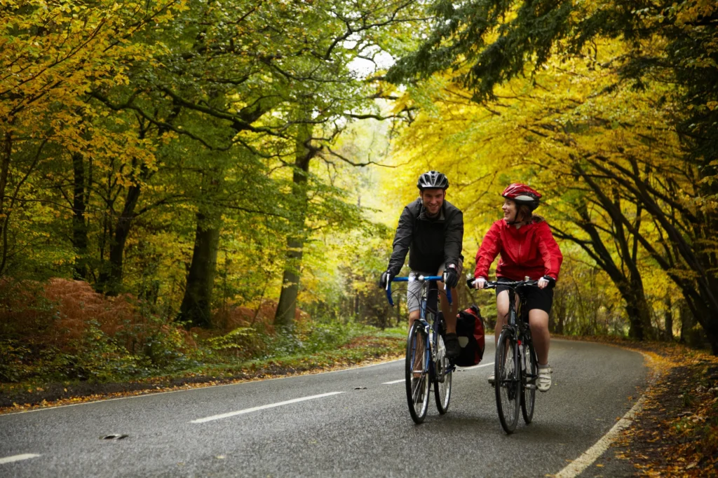 Due ciclisti in sella su una strada asfaltata circondata da alberi in autunno con fogliame giallo e verde.