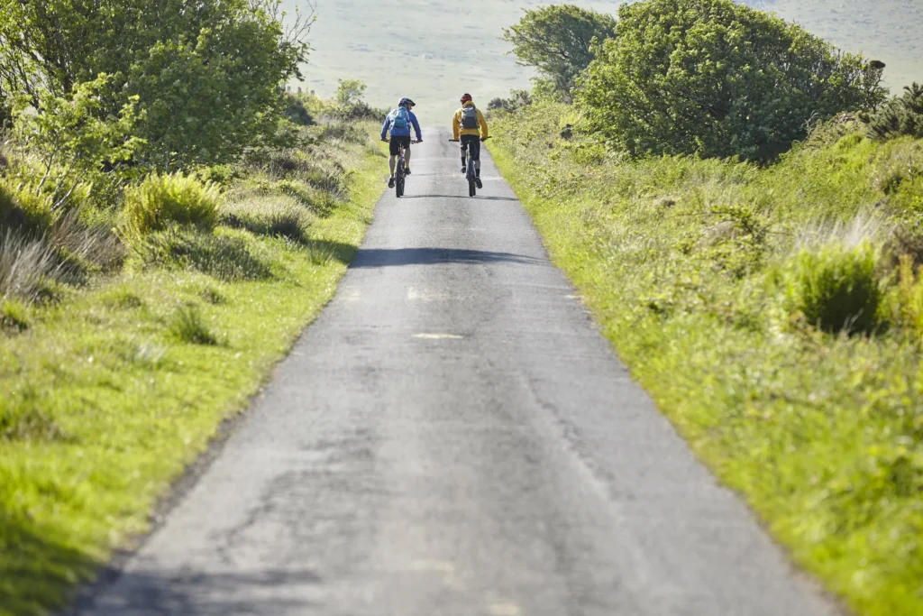 Due ciclisti su strada asfaltata circondata da vegetazione verde lussureggiante.