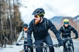 Gruppo di ciclisti in abbigliamento invernale pedala su strada con montagne e boschi sullo sfondo.