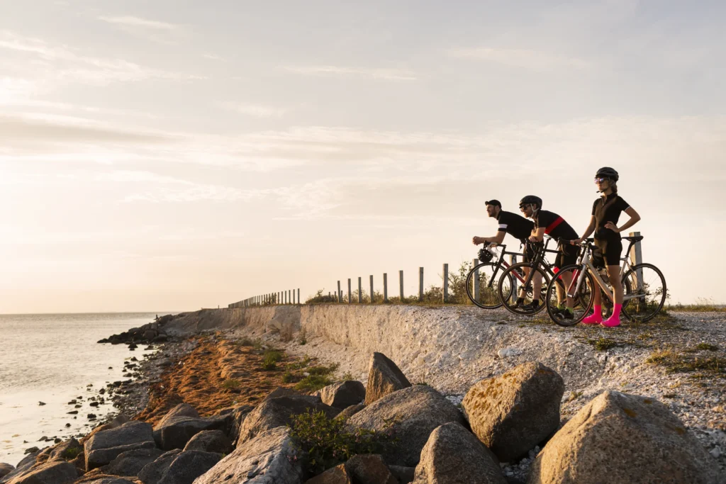 Tre ciclisti con le bici su una spiaggia di ciottoli affacciata al mare, con una staccionata sullo sfondo.