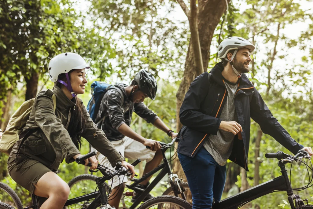 Tre ciclisti in casco pedalano insieme su un sentiero boscoso circondati da alberi e vegetazione verde.