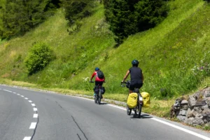 Due cicloturisti in salita su strada asfaltata circondata da prati verdi e boschi.