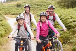 Famiglia di quattro persone in sella a mountain bike su sterrato in montagna, circondata da prati verdi e boschi.