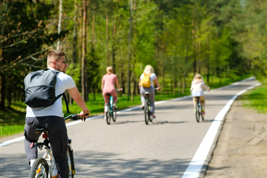 Ciclisti di diverse età pedalano su una ciclovia asfaltata circondata da bosco fitto di conifere.