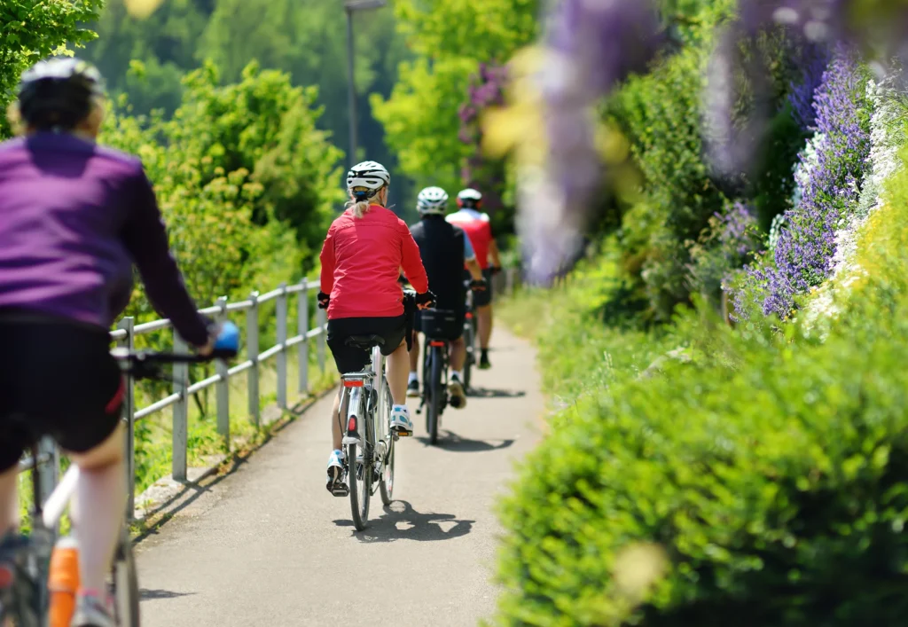 Gruppo di ciclisti su percorso pavimentato circondato da verde lussureggiante e fiori viola.