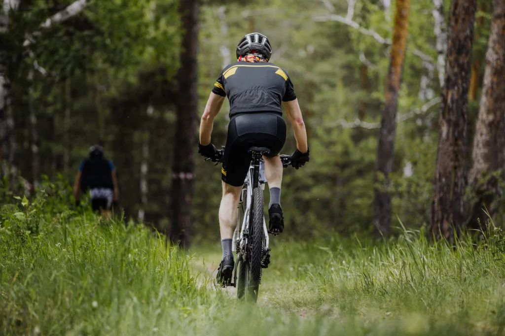 Ciclista in maglia nera e gialla pedala su sterrato tra alberi e erba verde in bosco.