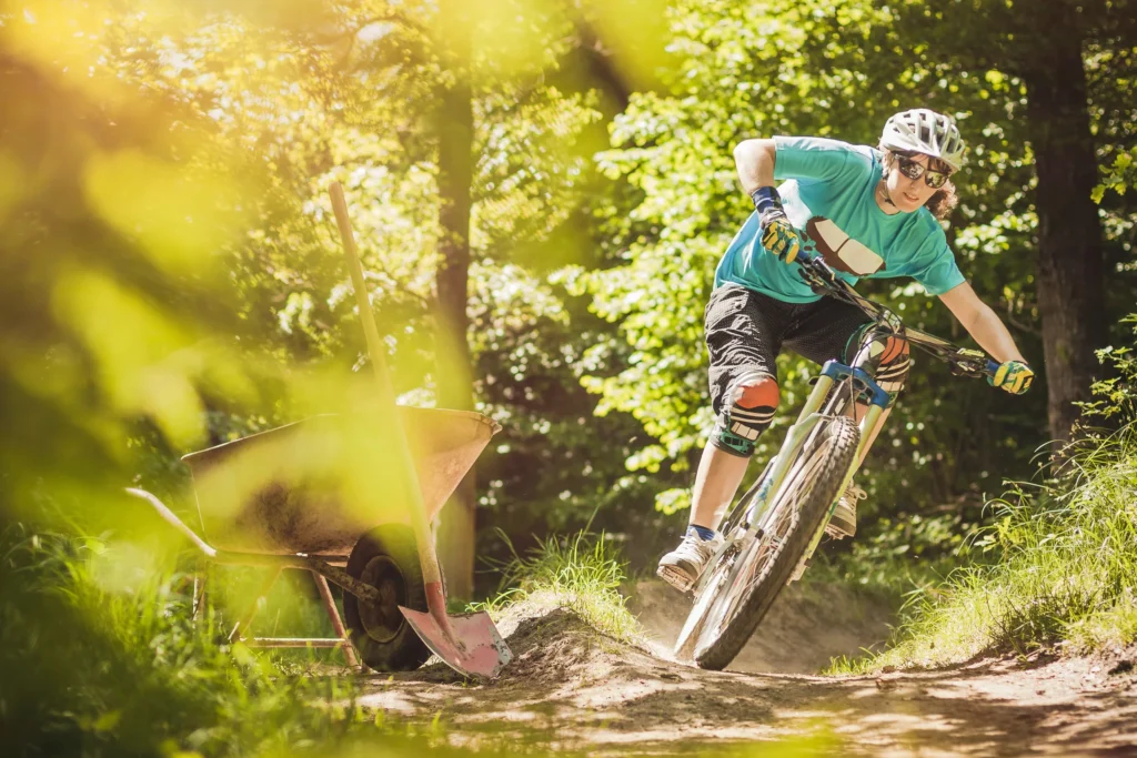 Mountain biker in green shirt rides a singletrack forest trail in bright sunlight, with old farm equipment visible on the side.