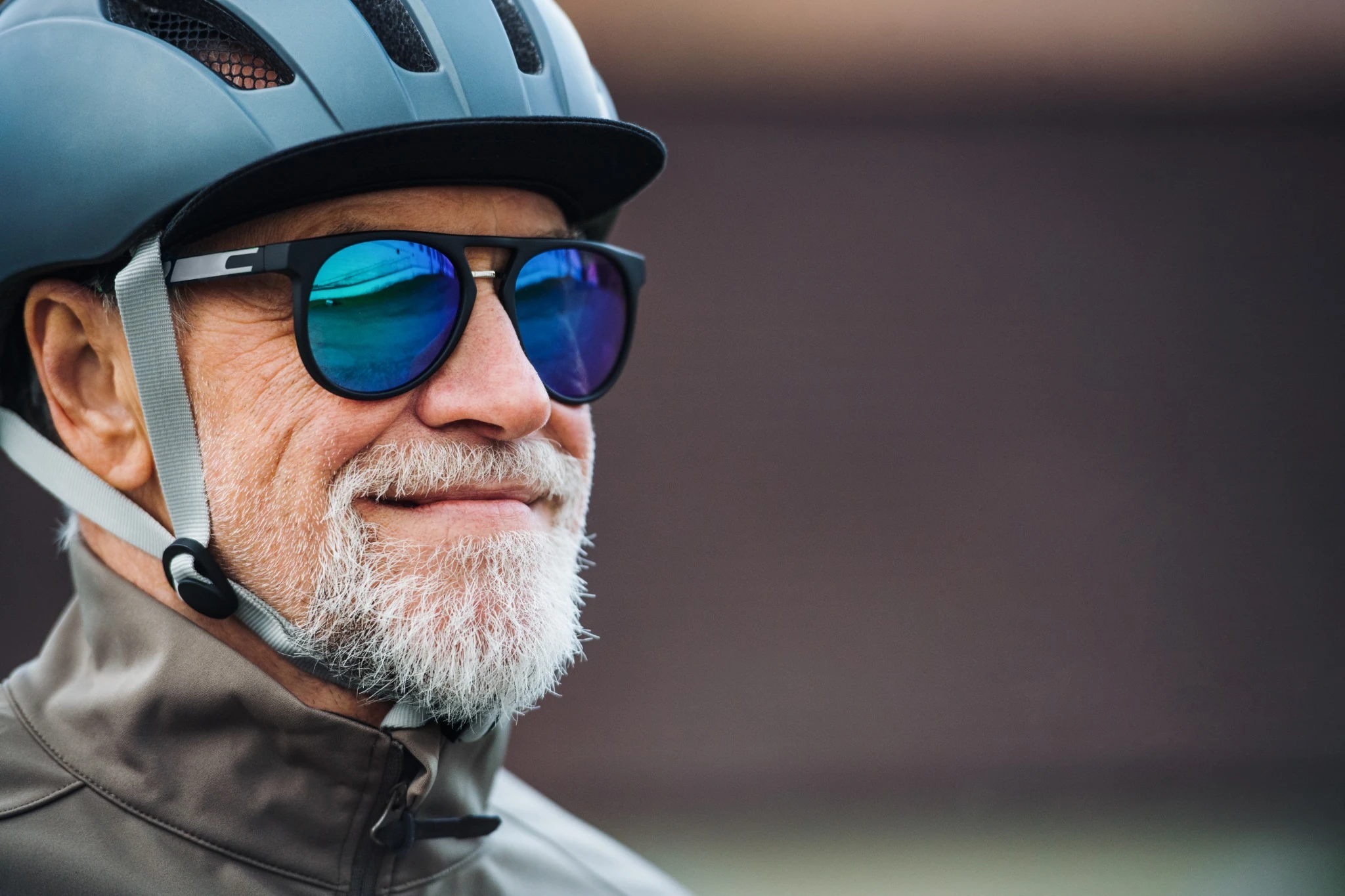 Ciclista con casco azzurro, occhiali da sole a lenti blu e barba bianca in primo piano.