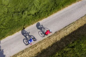 Due ciclisti su strada asfaltata stretta tra campi verdi, vista aerea con ombre nitide.