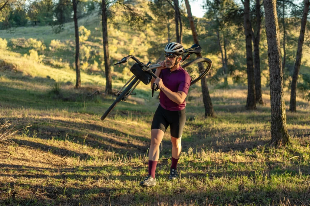 Ciclista con bicicletta in spalla cammina su sentiero nel bosco in luce dorata.